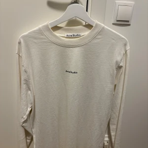 Vit långärmad tröja Acne Studios - Minimalistisk vit långärmad tröja från Acne Studios med diskret logga på bröstet. Tröjan har rund halsringning och är tillverkad i mjuk bomull, perfekt för en clean och stilren look.