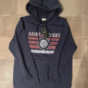 Marinblå hoodie från Saint Laurent - Säljer en marinblå hoodie från Saint Laurent med tryck i vitt och rött framtill. Tröjan har huva med snörning. Storlek L och nytt skick. Mitt pris 1900, kan diskuteras!!