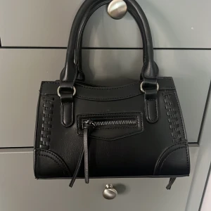 Svart handväska - Snygg svart handväska. Väskan har  detaljer med dekorativa sömmar och en dragkedjeficka framtill. Perfekt storlek för det viktigaste. 