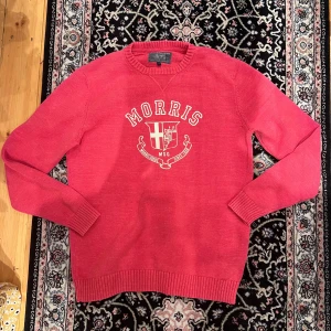Röd stickad tröja från Morris - Snygg rosa stickad tröja från Morris i 100% bomull. Tröjan har ett stort tryck med Morris-logga och emblem på bröstet, samt ribbade muddar vid hals, ärmslut och nederkant. Perfekt för dig som gillar klassisk college-stil och vill sticka ut med färg.