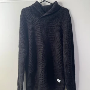 Svart stickad tröja från Jack & Jones - Mysig svart stickad tröja från Jack & Jones med grov struktur och hög krage. Perfekt för kyliga dagar och enkel att matcha med jeans eller chinos. Långärmad modell med avslappnad passform och ribbade muddar.