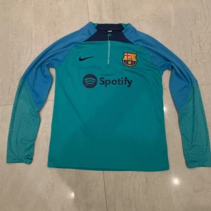 Blå långärmad FC Barcelona träningströja - Snygg långärmad träningströja från FC Barcelona i blå och turkos nyans. Tröjan har halv dragkedja, klubbmärke på bröstet och Nike-logga. Materialet är lätt och andas, perfekt för fotbollsträning. Spotify-logga på framsidan.