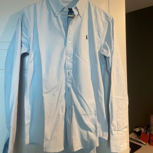 Ljusblå skjorta från Polo Ralph Lauren - Klassisk ljusblå skjorta från Polo Ralph Lauren med broderad svart logga på bröstet. Skjortan har button-down krage, långa ärmar och knäppning framtill. Tillverkad i mjuk bomull som känns skön mot huden. Perfekt för dig som gillar stilren och tidlös design. Prislapp är kvar och den skickas i orginalförpackning.