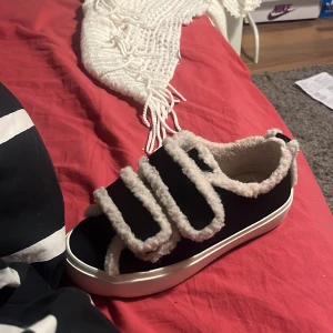 Svarta sneakers med fuskpälsdetaljer - Unika svarta sneakers med vit platt sula och fluffiga fuskpälsdetaljer längs kanterna och på de breda kardborrebanden. Skorna har rund tå och en mysig vibe, perfekta för dig som vill sticka ut med din stil. Köpt från inuikii