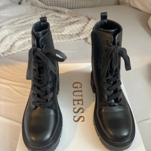 Svarta boots från Guess i skinn - Snygga svarta boots från Guess i skinn med grov sula och snörning framtill. Skorna har dragkedja på sidan för enkel påtagning och präglade Guess-loggor på utsidan. Perfekta för dig som vill ha en edgy look till höst och vinter.