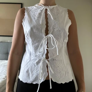 Vit broderad blus med knyt - Superfin vit blus med broderade blommor och knyt framtill. Ärmlös modell med vågiga kanter. Aldrig använd, då jag inte gillat modellen. Tryck inte på KÖP DIREKT utan skriv privat om du är intresserad! 💛