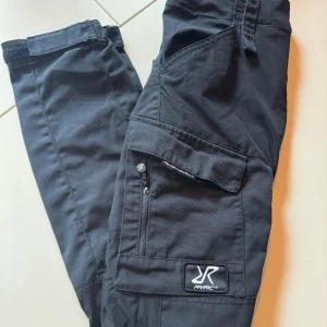 Svarta friluftsbyxor från RevolutionRace - Svarta friluftsbyxor från RevolutionRace med flera praktiska fickor, dragkedjedetaljer och logga på benet. Byxorna har elastisk midja och är tillverkade i slitstarkt material, perfekta för äventyr och outdoor. Snygg och funktionell design för en aktiv livsstil. Så fina på! 