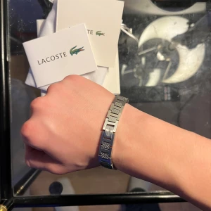 Lacoste silver armband  - Säljer en stilren Lacoste silverarmband  i originalförpackning. Armbandet levereras i en vit box med grönt Lacoste-logga och en vit tygpåse med snörning, också med den klassiska krokodilen. Perfekt accessoar för dig som gillar sportig och clean design.