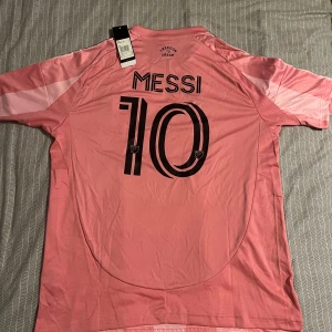 Lionel Messi  - Säljer en officiell Inter Miami CF fotbollströja från Adidas med Messi 10-tryck på ryggen. Tröjan är rosa med svarta detaljer, har korta ärmar, randigt mönster och klubbmärke på bröstet. Tillverkad i lätt och ventilerande AEROREADY-material.