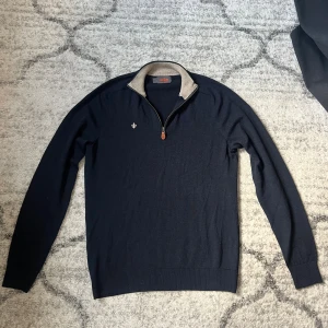 Mörkblå half zip tröja från Morris - Snygg mörkblå stickad tröja från Morris med half zip och kontrastfärgad krage i beige. Tröjan har lång ärm, ribbade muddar och en liten broderad logga på bröstet. Perfekt för lager-på-lager och stilren look.