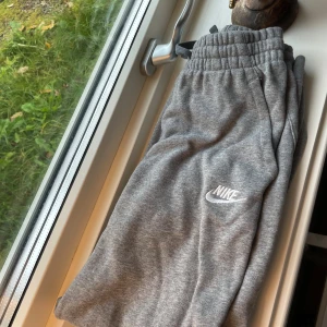 Grå mjukisbyxor från Nike - Grå mjukisbyxor från Nike med broderad vit logga på vänster ben. Byxorna har resår i midjan med snörning och är tillverkade i mjukt bomullsmaterial. Perfekta för chill eller träning, med klassisk rak passform och sidofickor.