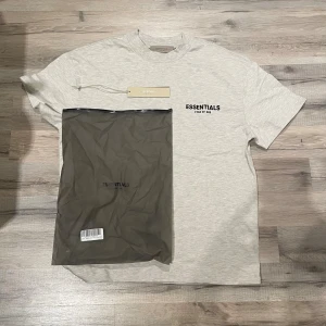 Grå Essentials Fear of God t-shirt - Snygg grå t-shirt från Essentials Fear of God med svart logotyptryck både fram och bak. Klassisk rund hals och korta ärmar. Tillverkad i mjuk bomull för skön passform. Perfekt för dig som gillar streetwear och cleana vibes.