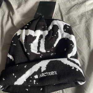 Svart och vit mössa från Arc'teryx - Snygg svart mössa från Arc'teryx med vit abstrakt grafik och broderad logga framtill. Mössan har uppvikt kant och är tillverkad i mjukt stickat material som håller dig varm. Perfekt för dig som gillar streetwear och vill sticka ut med cool design.