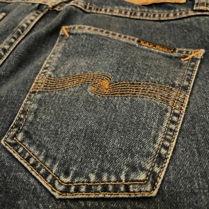 Blå jeans från Nudie Jeans 29/34 - Säljer ett par klassiska blå jeans från Nudie Jeans i storlek 29/34. Jeansen har raka ben, snygga kontrastsömmar och Nudie Jeans ikoniska broderi på bakfickorna. Tillverkade i slitstark bomull med en tidlös look som passar till allt.  Skick 8,5/10