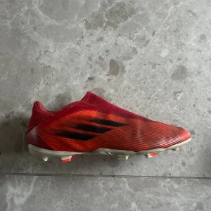 Röda Adidas fotbollsskor X - Säljer ett par röda Adidas X fotbollsskor med svart detalj på sidan och vit sula med röda dobbar. Skorna har en strömlinjeformad design utan snörning och är tillverkade i syntetmaterial för lätthet och bra grepp på planen.