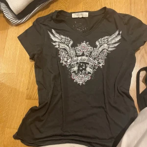 Svart t-shirt med vingar och rosor - Svart t-shirt med v-ringning från Black Lace Riders. Framsidan har ett stort tryck med vingar, pistoler och rosor i grått och vitt, samt texten 'Black Lace Riders'. Skön passform och cool bikerstil.