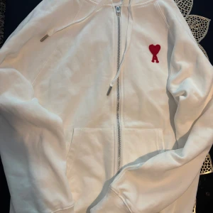 Vit hoodie med rött hjärta från Ami - Vit hoodie från Ami Alexandre Mattiussi med dragkedja, huva och snörning. På bröstet finns ett rött hjärta med bokstaven A. Tillverkad i mjuk bomull och har praktiska fickor framtill. Perfekt för en chill och stilren look.