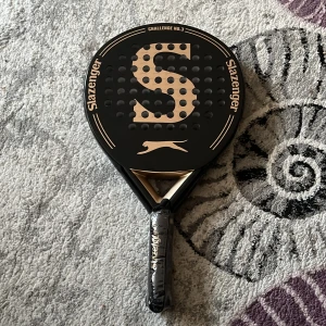 Challenge No.3 Padelracket - Slazenger Challenge No.3 padelracket i nyskick. Snygg svart och guld design, stort S på träffytan och originalplast kvar på handtaget. Perfekt för både nybörjare och erfarna spelare. Inga synliga skador eller slitage. Helt ny. Org pris 2500kr