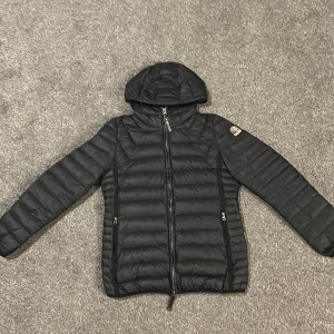 Svart dunjacka från Parajumpers L - Svart superlätt dunjacka från Parajumpers med huva och dragkedja framtill. Jackan har quiltad design, två fickor med dragkedja och klassisk PJS-logga på ärmen. Insidan är ljusgrå med orange detaljer och meshficka. Perfekt för kyliga dagar.