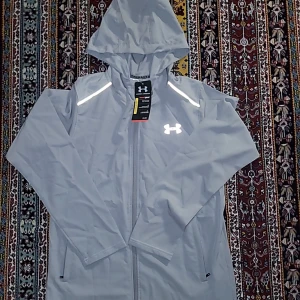 Grå vindjacka från Under Armour M - Snygg grå vindjacka från Under Armour med huva, hel dragkedja och reflekterande detaljer. Jackan har meshfoder, två sidofickor och logga på bröstet samt stor logga på ryggen. Perfekt för träning utomhus när det blåser. Fick den i present men tyvärr passar den inte mig