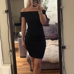 Svart offshoulder klänning - Snygg svart offshoulder klänning med tight passform och kort längd. Klänningen har knytband vid axlarna och är tillverkad i ett lätt rynkat material som ger en cool struktur. Perfekt för dig som gillar en stilren och trendig look 