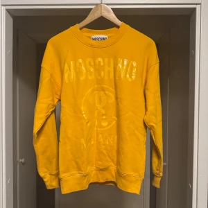 Gul Moschino tröja - Säljer en gul tröja från Moschino med glansigt tryck framtill. Tröjan har rund halsringning och långa ärmar. Perfekt statement-plagg för dig som gillar färg och vill sticka ut.