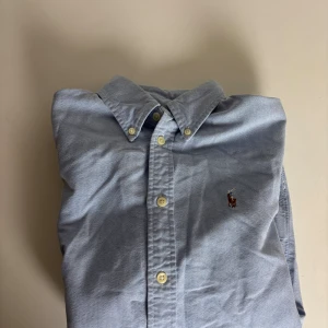 Blå skjorta från Ralph Lauren - Snygg ljusblå skjorta från Ralph Lauren med klassisk button-down krage och broderad logga på bröstet. Custom fit-modell med långa ärmar och vita knappar framtill. Skjortan är gjord i mjuk bomull och passar perfekt till jeans eller chinos.