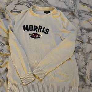 Vit stickad tröja från Morris - Vit stickad tröja från Morris med svart broderad logga och Union Jack-flagga framtill. Tröjan har rund hals, ribbade muddar och nederkant samt långärmad passform. Perfekt för dig som gillar klassisk stil med brittisk touch.