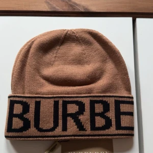 Beige stickad mössa från Burberry - Snygg beige stickad mössa från Burberry med bred uppvikt kant och stor svart logotyp runtom. Klassisk och stilren design som ger en exklusiv känsla. Perfekt accessoar för att lyfta din vinterstil.