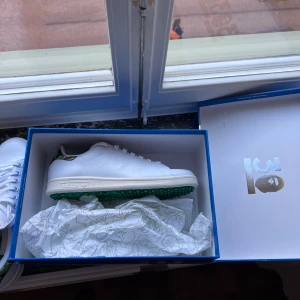 Golfskor Adidas Stan Smith x Bape sneakers - Helt nya Adidas Stan Smith x Bape sneakers i vitt skinn med gröna detaljer och camouflagemönster på hälen. Klassisk rund tå, snörning och grön gummisula med mönster. Ikonisk Stan Smith-tunga med grönt tryck. Levereras med originalkartong och taggar.