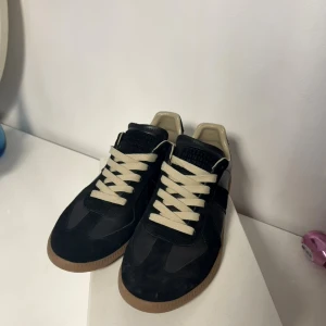 Svarta Maison Margiela sneakers - Maison Margiela sneakers i svart skinn och mocka med beige detaljer och snörning. Klassisk rund tå och platt gummisula i brunt. Pris kan diskuteras. Är du intresserad av fler bilder eller vill veta mer, är det bara att höra av dig!