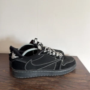 Travis Scott x Air Jordan 1 Retro Low - Säljer ett par svarta Nike Jordan 1 Low med tydliga vita kontrastsömmar och coola svartvita snören. Skorna har en klassisk siluett, rund tå och är gjorda i mocka med detaljer i skinn. Perfekta för dig som vill sticka ut med en unik sneaker.