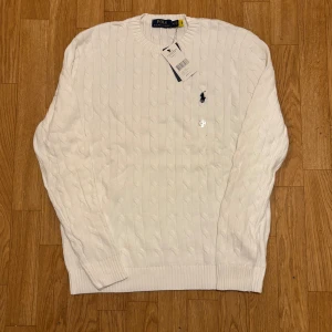 Vit kabelstickad tröja Polo Ralph Lauren - Snygg vit kabelstickad tröja från Polo Ralph Lauren i storlek M. Tröjan har rund hals, ribbade muddar och den klassiska broderade loggan i marinblått på bröstet. Perfekt för dig som gillar stilrena och tidlösa plagg med premiumkänsla. Extreeemt bra kvalite. Passa på och köp innan det tar slut