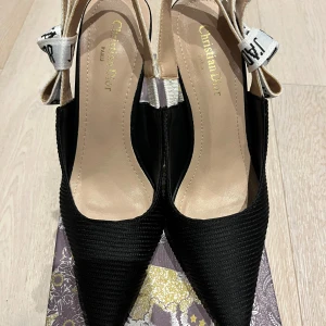 Svarta Dior J'Adior pumps med klack - Snygga svarta pumps från Dior med spetsig tå och hög klack. Skorna har ribbad textil på ovansidan och en vit rem med svart J'Adior-tryck och rosett bak. Insidan är beige och klacken är elegant smal. Perfekt för dig som gillar lyxiga detaljer.