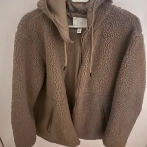 Beige teddyjacka med huva från H&M - Mysig beige teddyjacka från H&M med huva och dragsko. Jackan har dragkedja framtill och två fickor på sidorna. Perfekt för kyliga dagar och ger en avslappnad vibe.