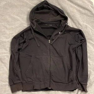 Svart hoodie från Under Armour - Svart hoodie från Under Armour med dragkedja framtill, huva med snörning och två stora fickor. Tröjan har en loose passform och är tillverkad i mjukt fleecematerial, perfekt för kyliga dagar. Klassisk och enkel design med ribbade muddar.