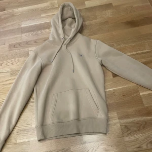 Beige hoodie från GANT med broderad logo - Säljer en beige hoodie från GANT i storlek XS. Tröjan har broderad GANT-logo på bröstet, klassisk känguruficka och snörning i huvan. Mjuk insida och ribbade muddar vid ärmslut och nederkant. Perfekt för chill dagar.