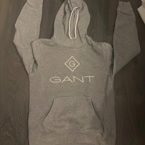 Grå hoodie från GANT med logga - Säljer en grå hoodie från GANT i storlek XS. Tröjan har vit dragsko i huvan, stor magficka och GANT-logga broderad framtill. Perfekt för chill dagar och enkel att matcha med jeans eller mjukisbyxor. 