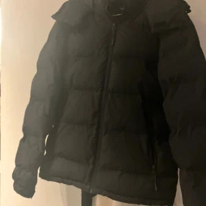 Svart pufferjacka med huva - Svart pufferjacka från Pull&Bear med huva och dragkedja framtill. Jackan är vadderad och har en klassisk, boxig passform med långa ärmar. Perfekt för kalla dagar när du vill hålla värmen och ändå se stilren ut.