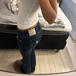 Mörkblå True Religion jeans bootcut - Säljer ett par mörkblå jeans från True Religion med klassiska kontrastsömmar och snygga fickdetaljer med lock och knappar bak. Modellen har låg midja och bootcut-ben, tillverkade i jeansmaterial. Perfekta för dig som gillar en tidlös och cool look. Tyvärr har de lite slitningar nere vid benen då dom har varit lite långa så har gått på dom