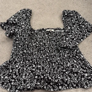 Blommig svartvit blus från H&M - Supersöt blommig blus från H&M i svart med vita blommor. Modellen har puffiga korta ärmar och smockad överdel som ger en snygg passform. Perfekt för dig som gillar romantiska vibes och vill sticka ut lite extra.
