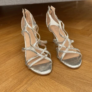 Silvriga klack-sandaler från Guess - Snygga silvriga sandaler från Guess med hög klack och ormskinnsmönster. Skorna har flera smala remmar, öppen tå och dragkedja bak med metall-detaljer. Perfekta för dig som vill sticka ut med ett glittrigt statement.