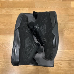  Air Jordan 4 Retro Black Cat - Nike Air Jordan 4 Retro Black Cat i helsvart färg med snygga meshpaneler och detaljer i mocka. Ikonisk Jumpman-logga på hälen och platt sula. Skorna har snörning och en chunky siluett som ger en cool streetwear vibe. Perfekt för dig som gillar klassiska sneakers med attityd. Priset är inte hugget i sten!