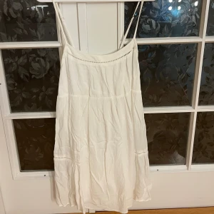 Vit sommarklänning med tunna axelband - Säljer en luftig och söt vit klänning med tunna axelband och avslappnad passform. Klänningen har en flowy siluett och fina detaljer med små hålbroderier. Perfekt för varma dagar och enkel att styla med sandaler eller sneakers. Köpt från PULL&BEAR.