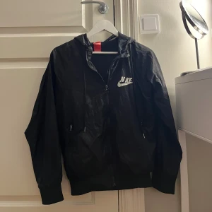 Svart Nike vindjacka med huva - Svart vindjacka från Nike med vit logga på bröstet. Jackan har huva med snörning, två fickor med dragkedja och ribbade muddar vid ärmslut och nederkant. Materialet är lätt och syntetiskt, perfekt för blåsiga dagar. Snygg och sportig look med hel dragkedja framtill. Den har ett litet hål på sidan men som går lätt att fixa med en lapp eller sy🤗 Den saknar även ett I i NIKE 💞