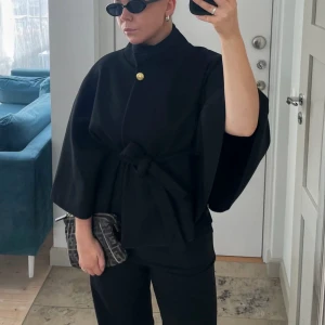 Svart kappa med bälte från Missay - Svart kappa/ poncho med vid passform och knytbälte i midjan. Jackan har en hög krage, vida ärmar och en snygg guldig knapp upptill.  