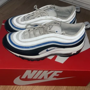 Nike Air Max 97 vit/blå/svart sneakers - Nike Air Max 97 sneakers med vågiga linjer och synlig Air-sula. Färgerna är vit, blå och svart med grå mesh och skinnpaneler. Rund tå, snörning och platt sula. Snygg retrostil med blå detaljer och reflekterande inslag. Perfekt för dig som gillar klassiska streetwear-vibbar.