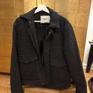 Rutig overshirt från These Glory Days - Snygg jacka i mörkgrått och svart hundtandsmönster från These Glory Days. Jackan har klassisk krage, två stora fickor framtill och dragkedja. Materialet är ullblandning med quiltat innerfoder, perfekt för kyliga dagar.