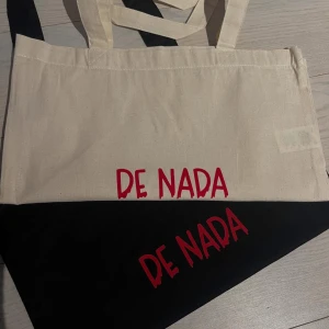 Shoppingväska med De Nada-tryck - Snygg shoppingväska i beige och svart med coolt rött De Nada-tryck på framsidan. Väskan har långa handtag och är tillverkad i slitstark bomull, perfekt för att bära med sig allt man behöver under dagen. Inspirerad av Alexander ernstbergers aura och grisch-trend. Helt nya och egentryckta!