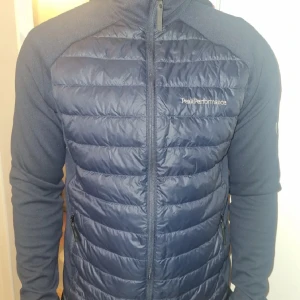 Mörkblå cardigan Peak Performance - Mörkblå cardigan från Peak Performance med quiltad framsida och rygg, huva och dragkedja. Jackan har släta ärmar i mjukt material och broderad logga på bröstet. Perfekt för lite kyliga dagar när du vill ha både stil och komfort. Fast pris.